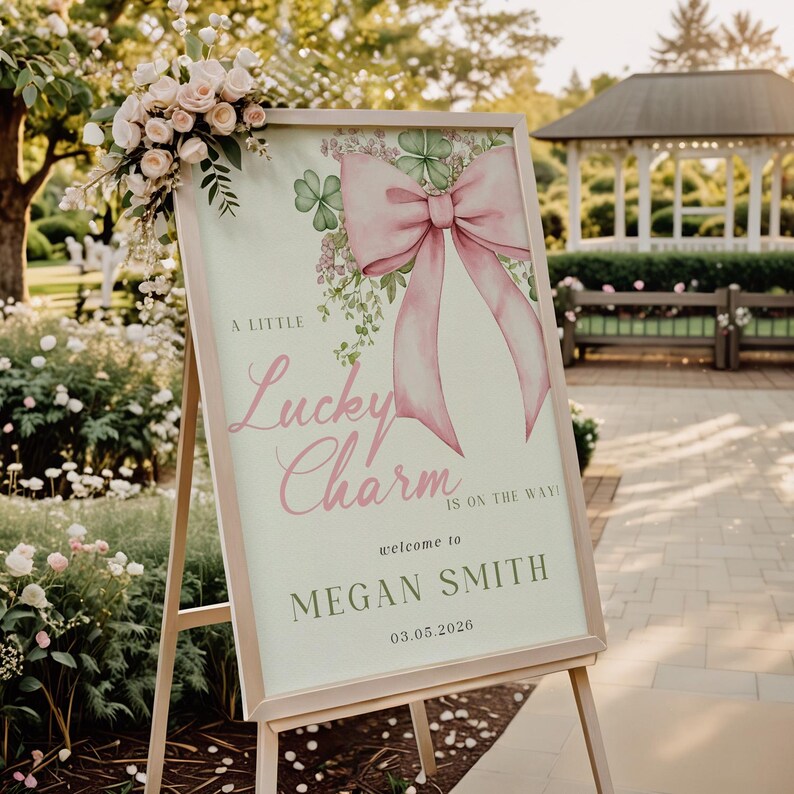 May include: A wooden easel displays a sign with a pink bow and the words "A Little Lucky Charm is on the way!" The sign welcomes Megan Smith and includes the date 03.05.2026. The sign is decorated with shamrocks and floral accents.