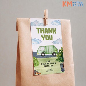 May include: A gift tag with a watercolor illustration of a green garbage truck, clouds, and grass. The tag reads "THANK YOU" and "FOR CELEBRATING WITH US". Includes the name "NATHANIEL". Attached to a brown paper bag with a wooden clip.