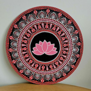 May include: A round wooden mandala with a pink lotus flower design in the center. The mandala is painted with black, white, pink, and red dots.