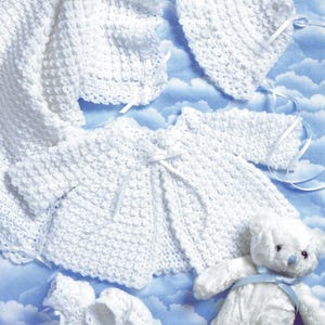 Puede incluir: Un conjunto de bebé de ganchillo blanco, que incluye una manta, un gorro, una chaqueta, patucos y un osito de peluche con una cinta azul. El conjunto está hecho de un tejido abierto texturizado. Cintas blancas acentúan la chaqueta y el gorro. El fondo es un cielo azul con nubes.