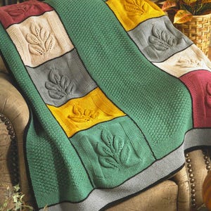 Puede incluir: Una manta tejida con un diseño de hojas. La manta presenta cuadrados verdes, amarillos, grises, crema y burdeos, cada uno con un diseño de hojas en relieve. La manta está sobre una silla.