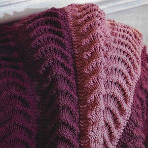 May include: A knitted throw blanket in shades of purple and pink. The blanket is made with a textured stitch that creates a wave-like pattern.