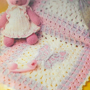 May include: A crocheted baby blanket in shades of pink, white and lavender with a crocheted teddy bear wearing a dress on top of it.