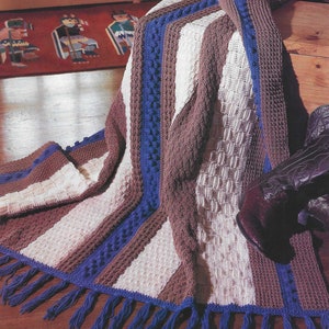 May include: A crocheted throw blanket with a striped pattern in brown, white and blue. The blanket has a fringe border and is draped over a wooden surface.
