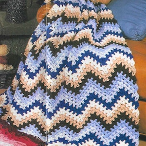 Op de afbeelding: Een gehaakt deken met een chevron-patroon in tinten blauw, bruin, wit en beige. Het deken is gedrapeerd over een bruine leren stoel.