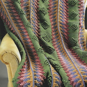 May include: A handmade, textured blanket in shades of green, pink, blue, and gold. The blanket features a repeating pattern of diamond shapes and textured borders, draped over a yellow armchair.