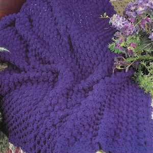 Op de afbeelding: Een paarse gehaakte deken met een textuur, een bobbelig ontwerp. De deken is gedrapeerd over een bank met een stoffering met bloemenmotief.