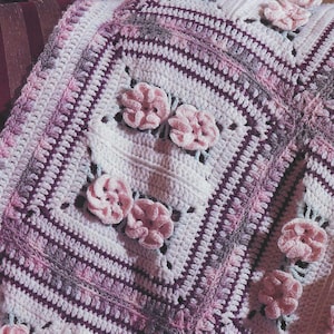 Könnte beinhalten: Eine weiße Häkeldecke mit einem rosa und violetten Rand. Die Decke hat ein sich wiederholendes Muster aus rosa gehäkelten Blumen.