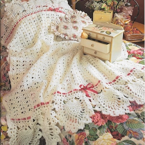 May include: A white crocheted blanket with a scalloped edge and pink ribbon trim. The blanket is draped over a bed with a floral patterned bedspread.