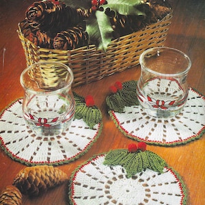 Puede incluir: Tres posavasos de ganchillo blanco con ribetes rojos y verdes. Cada posavasos presenta una hoja de acebo verde con bayas rojas en el centro.
