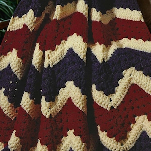 May include: A handmade crochet blanket with a chevron pattern. The blanket features alternating rows of cream, burgundy, and navy blue. The textured stitches create a cozy and inviting look, perfect for home decor.