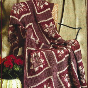 May include: A burgundy and cream-colored crocheted blanket draped over a black metal chair. The blanket features a geometric pattern with square motifs and a textured appearance. The chair is positioned near a potted plant with red flowers.
