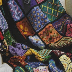 Könnte beinhalten: Eine farbenfrohe, handgefertigte Patchworkdecke. Die Decke zeigt eine Vielzahl geometrischer Muster und Designs in verschiedenen Farben, darunter Blau, Orange, Grün und Lila. Die Quadrate sind schwarz umrandet, was einen auffälligen Kontrast erzeugt.