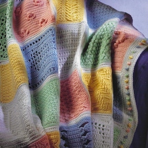 May include: A colorful, handmade crochet blanket. The blanket features a patchwork design with squares of various colors, including pink, yellow, blue, and green. Different crochet stitches create textured patterns. The blanket is draped, showcasing its soft and cozy appearance.