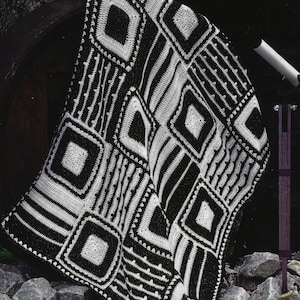 May include: A black and white crocheted blanket with geometric patterns, including squares and diamonds. The blanket is draped over a dark surface and rests on a bed of rocks. The design features bold lines and shapes.