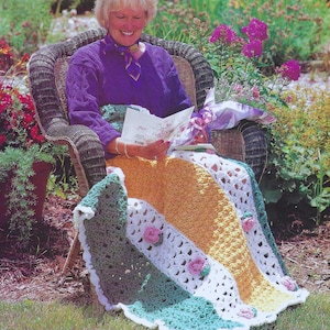 Pode incluir: Uma mulher está sentada em uma cadeira de vime em um jardim, envolvida em um cobertor de crochê com listras verdes, brancas, amarelas e rosas. O cobertor tem flores de crochê nos cantos.