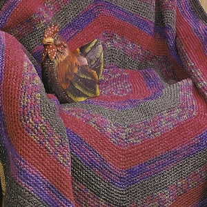 Puede incluir: Una manta tejida con un patrón geométrico en tonos de rojo, morado y gris. Una figura decorativa de gallo de madera descansa sobre la manta. La manta está doblada sobre una silla.
