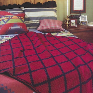 May include: A red and navy blue crocheted throw blanket with a checkered pattern. The blanket is draped over a bed with a wooden headboard and colourful pillows.