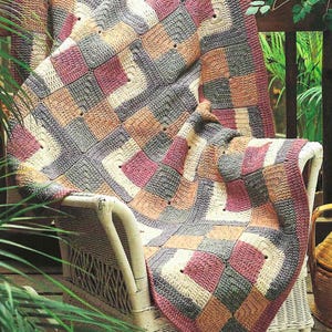 May include: A crocheted throw blanket with a patchwork design in shades of pink, beige, grey, and white. The blanket is draped over a white wicker chair on a wooden porch.