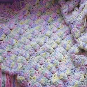 Puede incluir: Una manta de bebé de ganchillo en colores pastel de rosa, amarillo, verde y morado. La manta tiene un diseño texturizado y abultado y un borde de flecos.