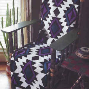 Könnte beinhalten: Eine gehäkelte Wolldecke mit einem geometrischen Muster in den Farben Lila, Blau, Schwarz und Weiß. Die Decke liegt auf einem Holzstuhl mit grünem Sitz.