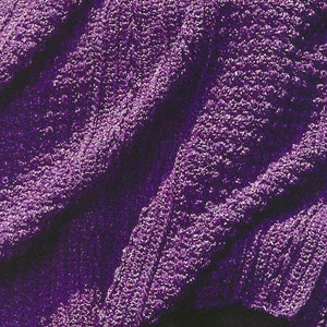 Puede incluir: Una manta de ganchillo de color morado con un patrón texturizado. La manta está doblada y drapeada sobre una pila de piedras.