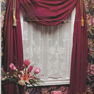 May include: A white crocheted curtain with a floral design hangs in a window with burgundy curtains and tassels. The window is framed by a white trim and the curtains are pulled back to reveal the crocheted curtain.