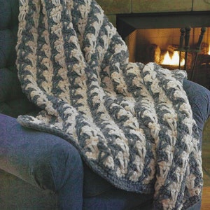 May include: A soft, cozy blanket with a striped pattern in shades of gray and white. The blanket is made of a fluffy yarn and is draped over a blue armchair.