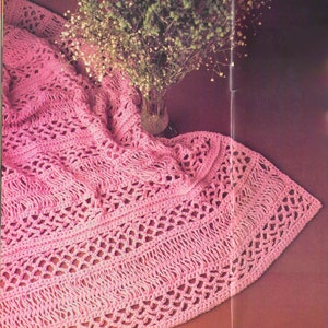 May include: A pink crocheted throw blanket with a delicate openwork pattern. The blanket is draped over a surface and has a soft, textured look.