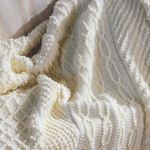 Pode incluir: Uma manta de crochê branca com um padrão texturizado e irregular. A manta está dobrada e drapeada sobre uma superfície marrom claro.