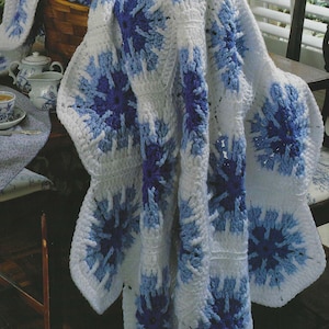 May include: A handmade crochet blanket in white, light blue, and dark blue. The blanket features a repeating floral pattern. The blanket is draped over a wooden table and chair, with a wicker basket and tea set in the background.