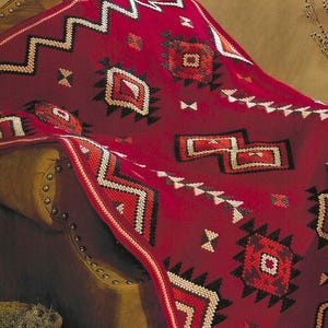 May include: A red blanket with a Southwestern design draped over a brown leather couch. The blanket features geometric patterns in white, black, and orange. The design includes zigzags, diamonds, and other shapes.