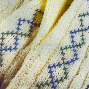 May include: A close-up of a crocheted blanket with a diamond pattern in blue and green on a cream background.