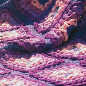 Puede incluir: Una manta de ganchillo con un diseño texturizado y abultado en tonos de morado y rosa.