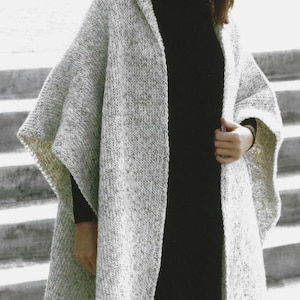 Può includere: Un mantello con cappuccio in tessuto a maglia grigio screziato. Il mantello è indossato sopra un dolcevita e un vestito neri. I bordi del mantello hanno un colore più chiaro, bianco sporco. Lo sfondo è una scala di cemento.