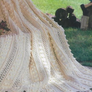 May include: A cream-coloured, hand-crocheted blanket with detailed patterns and a scalloped edge. The blanket has vertical textured stripes, openwork sections, and decorative bobbles. Tassels are attached to the bottom edge. The blanket is draped over a surface, suggesting a cosy, handmade home decor item.