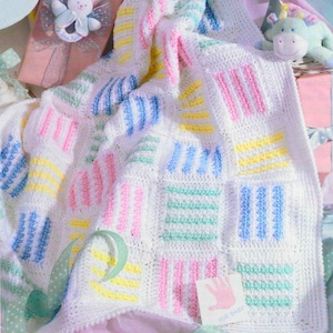 May include: A handmade, white crochet baby blanket with pastel stripes in pink, blue, yellow, and green. The blanket features a square pattern design. A small card with a pink handprint and the words "FOR BABY" is visible.