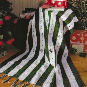 May include: A white and green crocheted throw blanket with a snowflake pattern. The blanket has fringe on the bottom edge and is draped over a table with wrapped gifts.