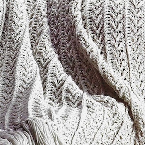 May include: A light gray, hand-crocheted blanket with a textured, chevron-like pattern. The blanket has a fringe along one edge. The soft, neutral color and intricate design suggest a cozy, handmade home decor item.