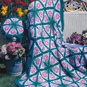 Op de afbeelding: Een gehaakt deken en kussen met een bloemenpatroon in tinten roze, wit en groen. Het deken is over een witte rieten stoel in een tuin gelegd.