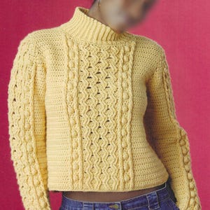 May include: A yellow crochet sweater with a high collar and long sleeves. The sweater features a textured pattern on the front and sleeves, with a ribbed collar and cuffs. The model is wearing blue jeans. The sweater is made of a soft, comfortable material.