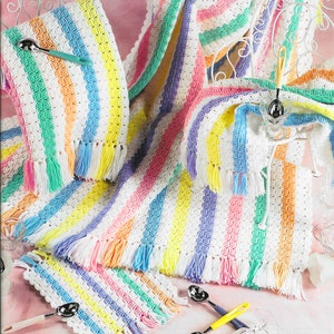 May include: A set of three crocheted striped blankets in white, pink, yellow, blue, and green with fringe trim. The blankets are arranged on a pink surface with silver ice cream scoops.