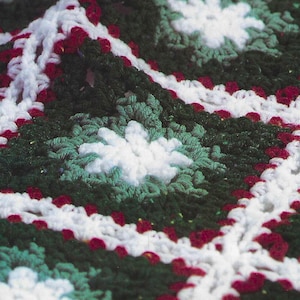 Könnte beinhalten: Eine gehäkelte Decke mit einem sich wiederholenden Muster aus weißen, grünen und roten Quadraten. Die Decke ist aus weichem, strukturiertem Garn gefertigt.