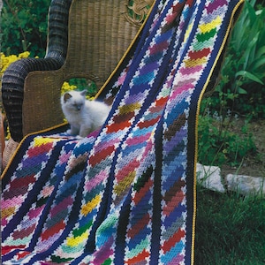 May include: A colorful crocheted blanket with a yellow border draped over a wicker chair. A white kitten sits on the blanket.