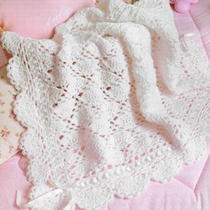 以下が含まれることがあります： 白いかぎ針編みのベビーブランケット。スカラップエッジとリボンが特徴です。ピンクのキルトの表面と「baby」の刺繍が入ったピンクの枕の上に掛けられています。ブランケットのオープンウィーブデザインが見えます。