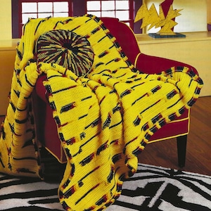 May include: A yellow crocheted throw blanket with a black and red trim. The blanket is draped over a red armchair with a colorful round pillow. The blanket has a repeating pattern of yellow and black stripes.