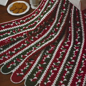 May include: A red, green, and white crocheted blanket with a scalloped edge. The blanket features alternating stripes of red and green, with white accents. The blanket is displayed with a plate of cookies, a mug of hot chocolate, and wrapped gifts.