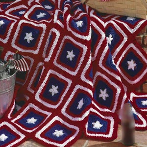 May include: A handmade crochet blanket in red, white, and blue, featuring a star pattern. The blanket is made of squares with a white star in the center of a blue square, surrounded by a white border and red background. The blanket is draped over a surface.