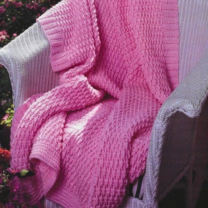 May include: A pink crocheted throw blanket draped over a white wicker chair. The blanket has a textured stitch pattern and is made with soft yarn.
