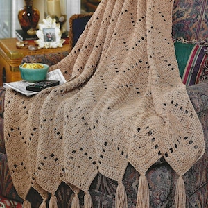 Puede incluir: Una manta de ganchillo beige con un diseño geométrico y borlas. La manta está sobre un sillón estampado. El diseño de ganchillo presenta cuadrados calados y un borde festoneado con borlas.